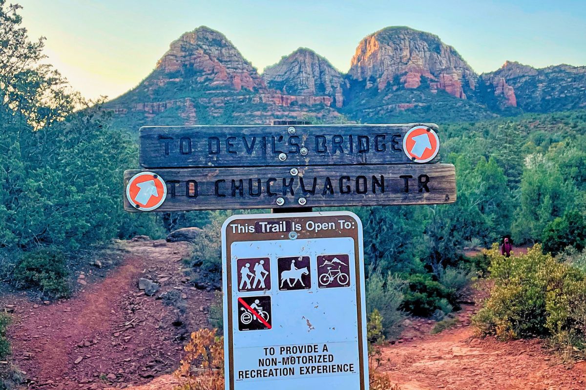 Trail sign pointing to Devil's Bridge and Chuck Wagon Trail in Sedona, with majestic red rock formations in the background.