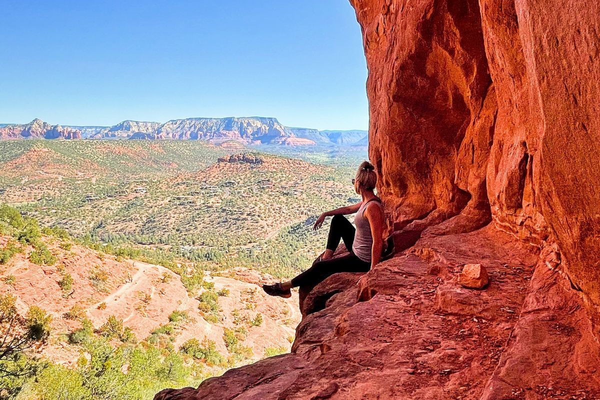 Kate sitting at the edge of a cave, admiring the expansive view of Sedona's red rock landscape, a must-see on any Sedona itinerary.