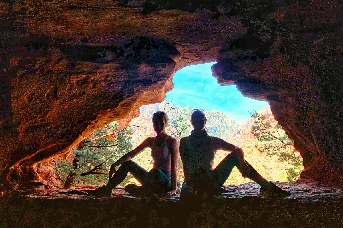 Kate and a friend sitting on a rock ledge with the view from the hidden Soldier Pass Cave on the Soldier Pass Trail in Sedona behind them.