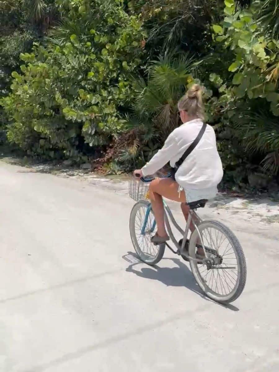 A woman riding a bicycle along a sandy path bordered by lush greenery in Caye Caulker, Belize. She is wearing a white shirt and carrying a shoulder bag.