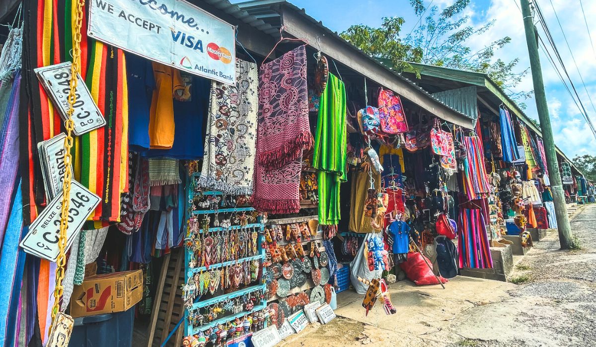 A bustling market stall in San Ignacio, Belize, displaying a vibrant array of colorful textiles, handcrafted jewelry, and souvenirs. License plates and various woven items hang alongside the stall, adding to the eclectic and lively atmosphere.