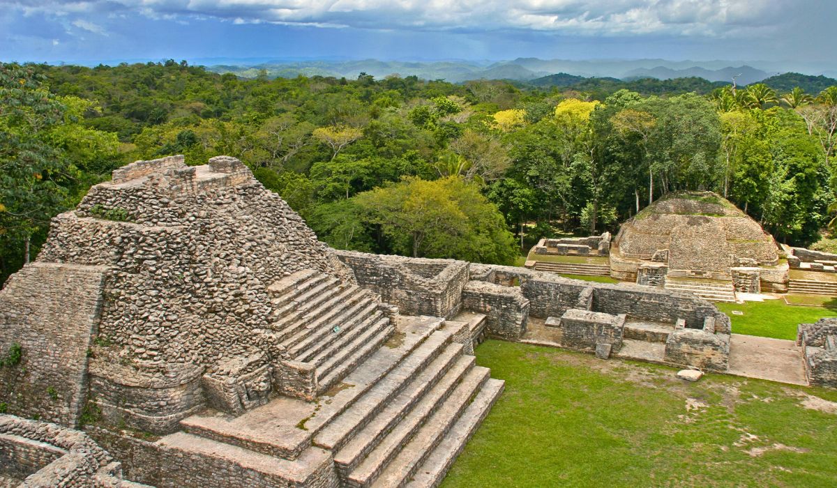 The ancient Mayan ruins of Caracol in Belize, featuring large stone pyramids and structures surrounded by dense jungle. The stepped pyramids rise prominently, showcasing the intricate stonework and historical significance of this archaeological site. The expansive green landscape extends into the distance under a cloudy sky.