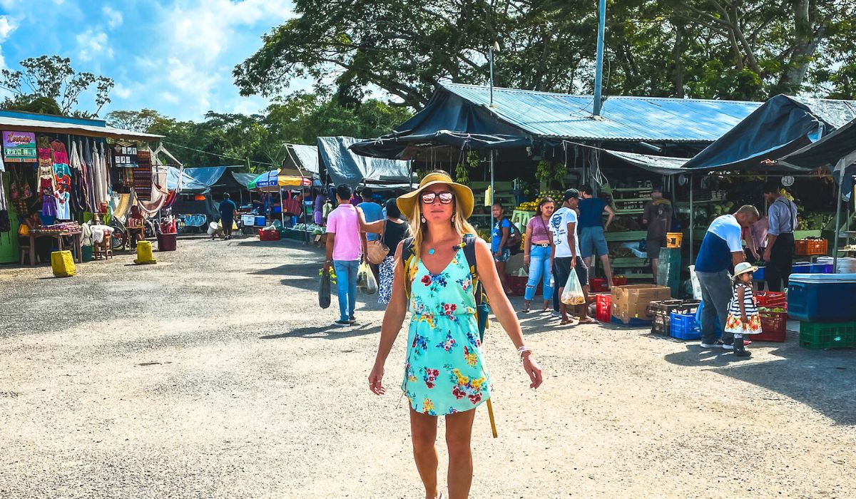 Kate stands in the middle of a bustling market in downtown San Ignacio, Belize, wearing a floral dress, sunglasses, and a sunhat. Various stalls line the street, displaying colorful textiles, fresh produce, and other goods. Shoppers and vendors engage in lively exchanges under the bright, sunny sky.