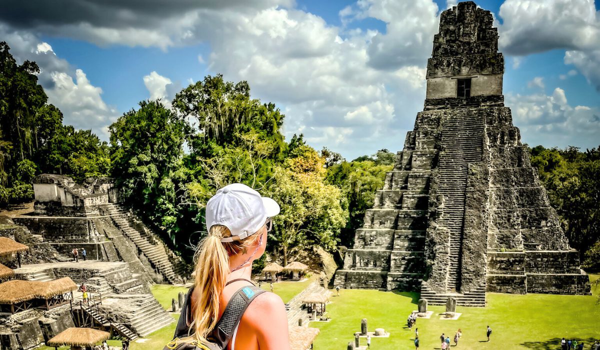 Kate, wearing a white cap and a backpack, gazes at the towering ancient pyramid of Tikal in Guatemala. The impressive stone structure rises majestically against a backdrop of lush green trees and a partly cloudy sky. Other visitors explore the archaeological site below.