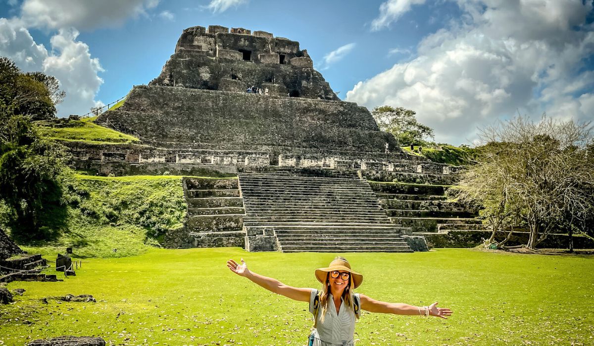 Kate's Crossing, wearing a hat and sunglasses, stands with her arms outstretched in front of the ancient Mayan ruins at Xunantunich in San Ignacio, Belize. The massive stone pyramid, featuring several tiers and a few trees surrounding it, towers impressively under a partly cloudy sky.