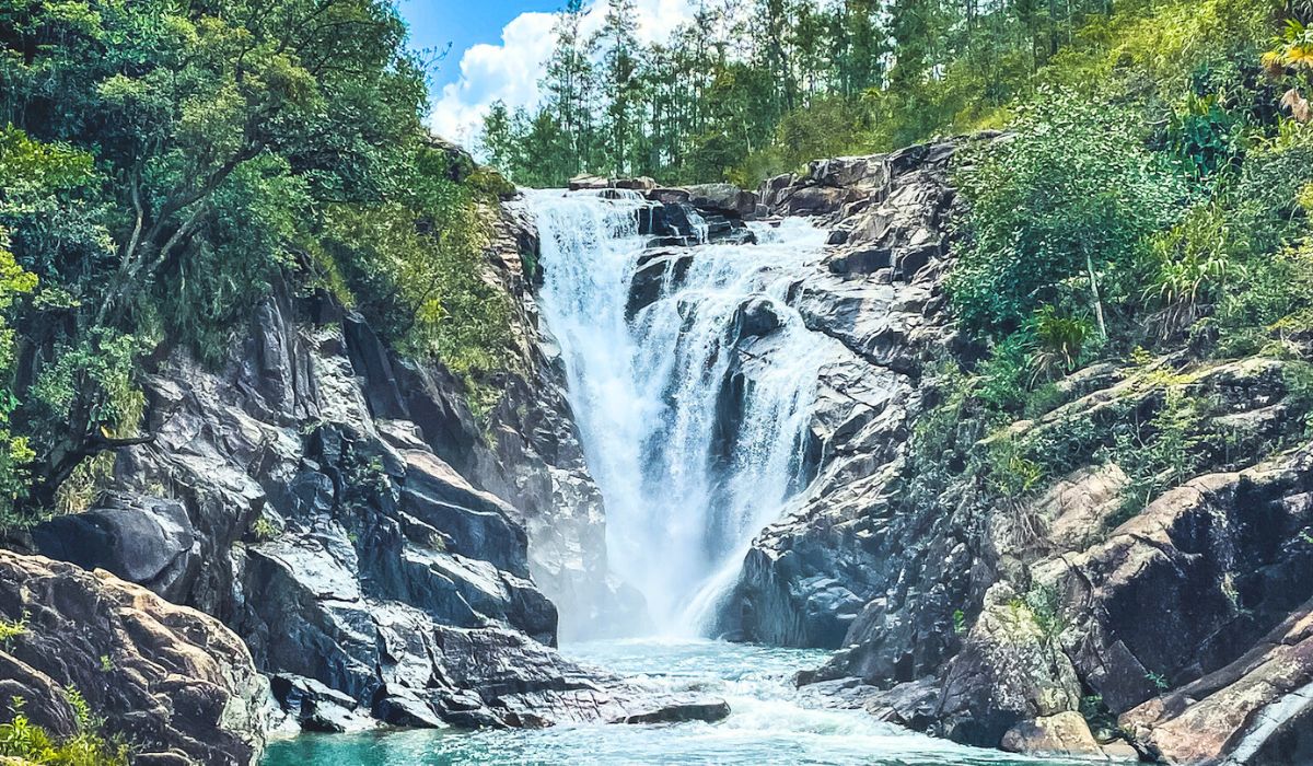 A majestic waterfall known as Big Rock Falls cascades down rugged, rocky cliffs into a turquoise pool below, surrounded by lush green foliage and trees under a bright blue sky.