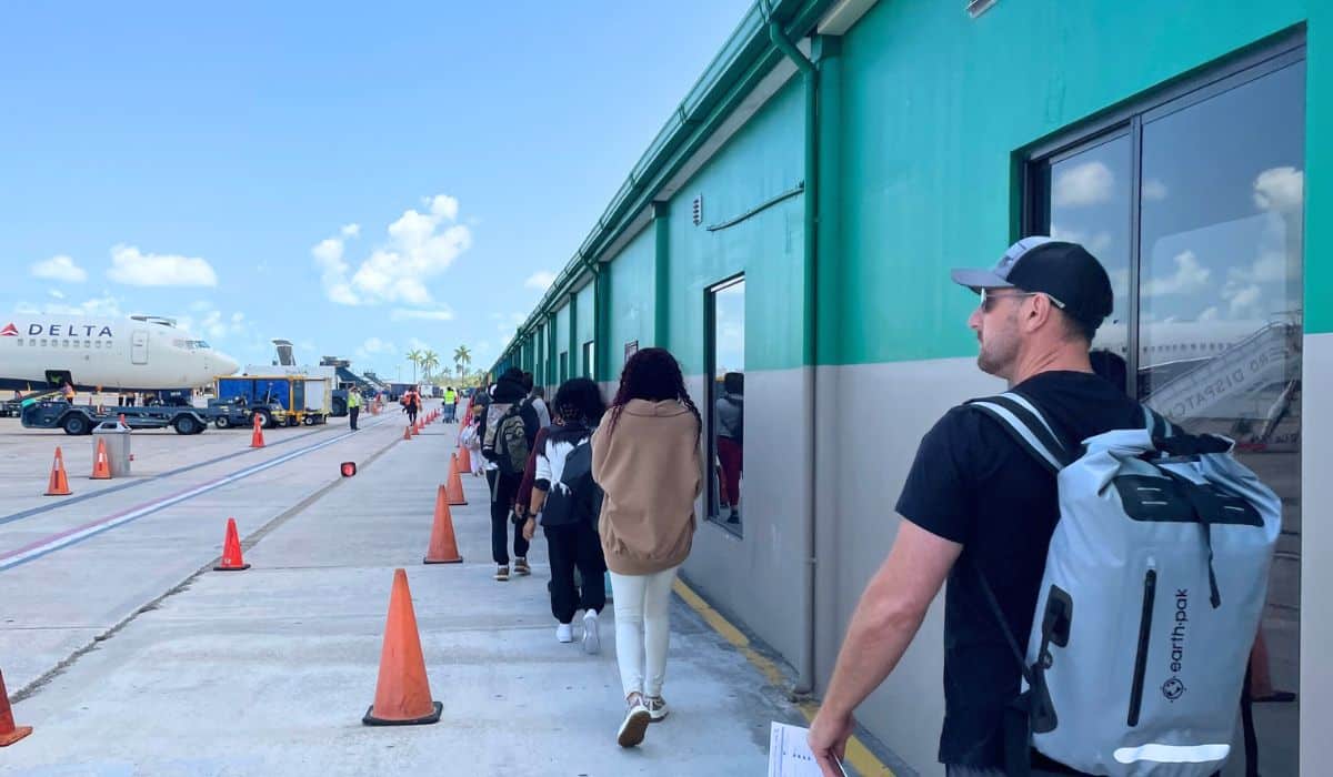 Passengers walk in a line along the tarmac towards the terminal building after landing at Belize International Airport in Belize City. A Delta airplane is parked nearby, and orange cones line the pathway under a bright, clear sky.