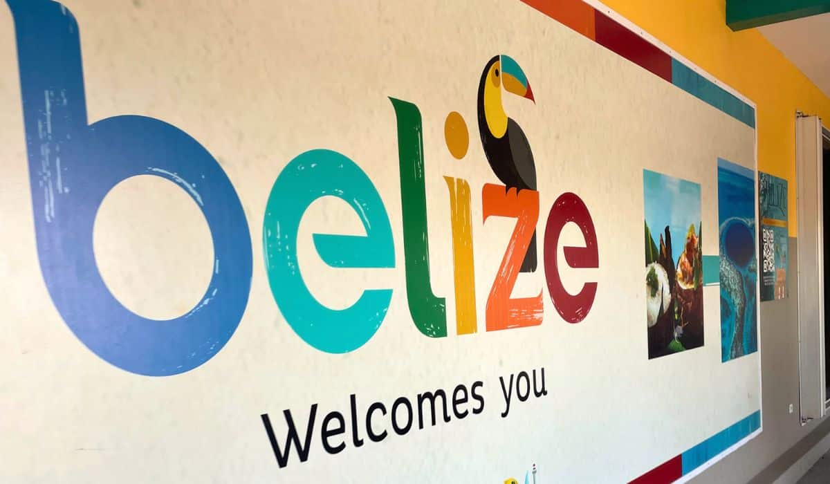 A colorful mural at the Belize City airport reads "Belize Welcomes You" with the word "Belize" featuring a toucan perched on the letter "i". The mural is adorned with vibrant images of Belizean landscapes and attractions, highlighting the natural beauty and cultural richness of the country.