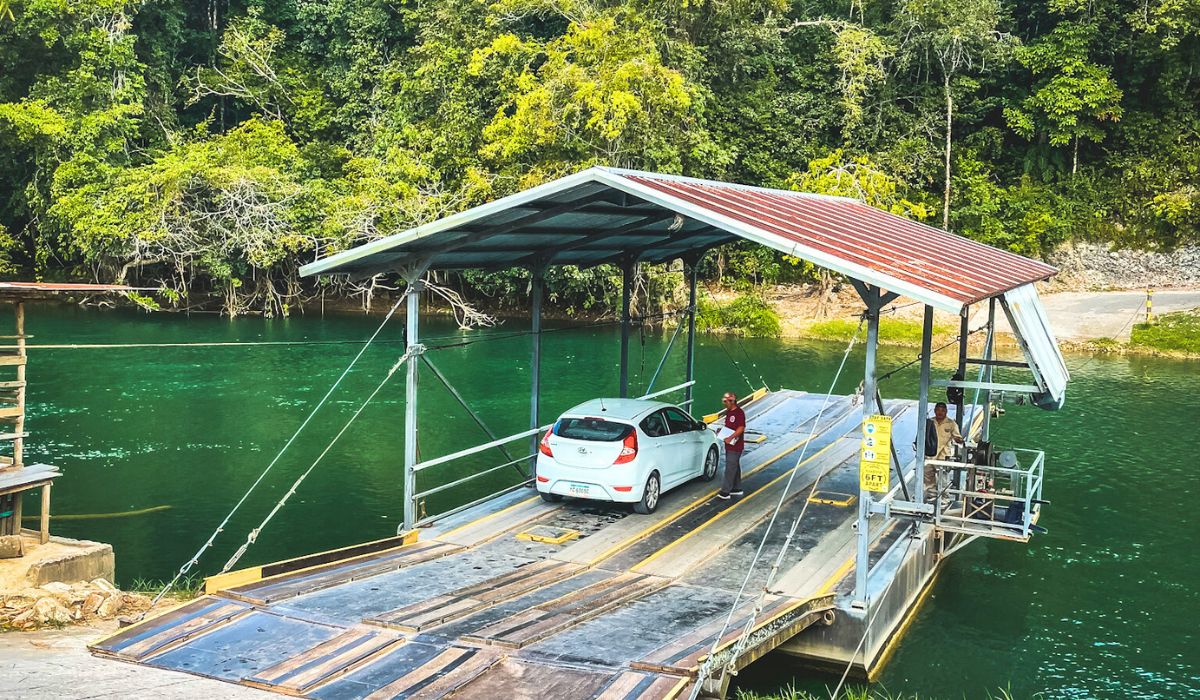 A white rental car is parked on a ferry as it crosses a serene river in San Ignacio, Belize. The ferry has a metal roof and is operated by a few attendants, surrounded by lush green trees and calm, emerald water.