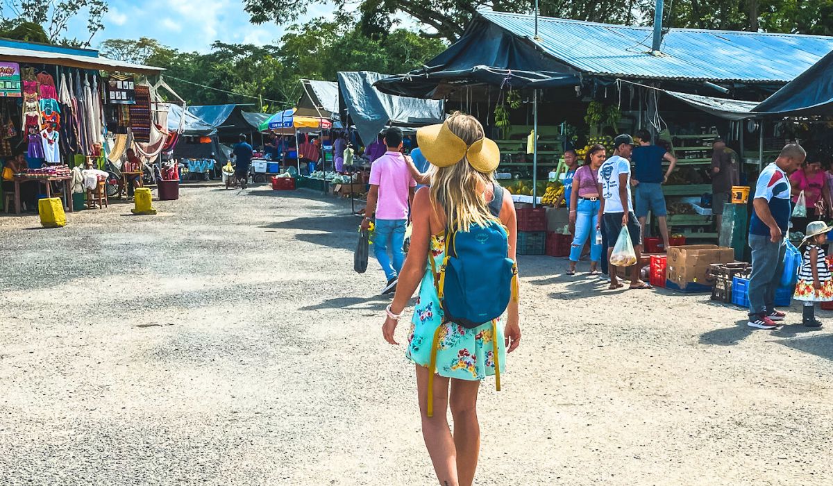 Kate from Kate's Crossing Blog, with a blue backpack and sun hat, walks through a bustling outdoor farmers market in San Ignacio, Belize. Stalls are filled with colorful textiles and fresh produce, capturing the vibrant local culture during her journey from Belize City to San Ignacio.