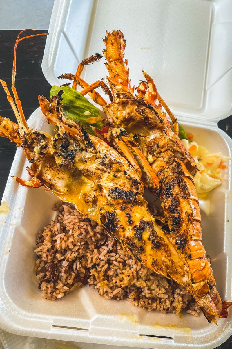 A takeout container filled with a serving of grilled lobster, seasoned and charred to perfection, accompanied by a side of rice and beans, and a small portion of coleslaw. The lobster is split open, revealing its succulent meat, while the rice and beans add a traditional Belizean touch to the meal.