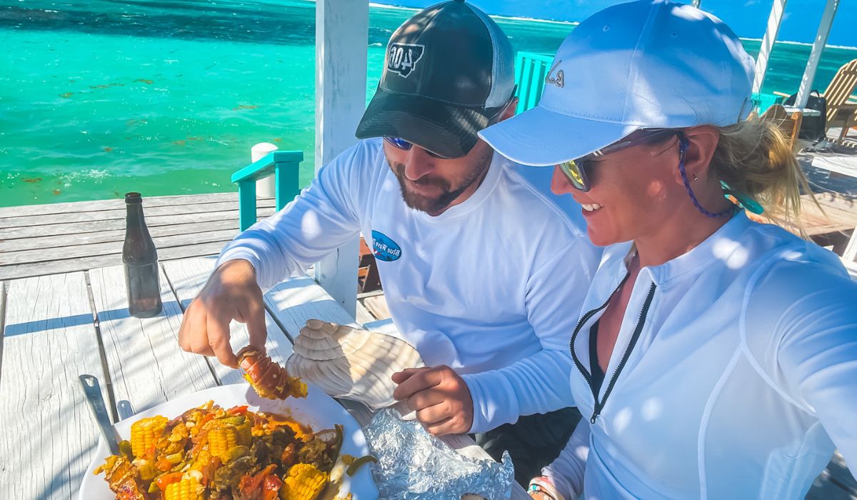 Kate and her husband enjoy a meal on a sunny dock overlooking the turquoise waters of Belize. The man is picking up a piece of seafood from a plate filled with Belizean foods, including corn on the cob and a variety of seasoned vegetables. Both are dressed in casual, sun-protective clothing and appear to be relishing their outdoor dining experience in this tropical paradise.