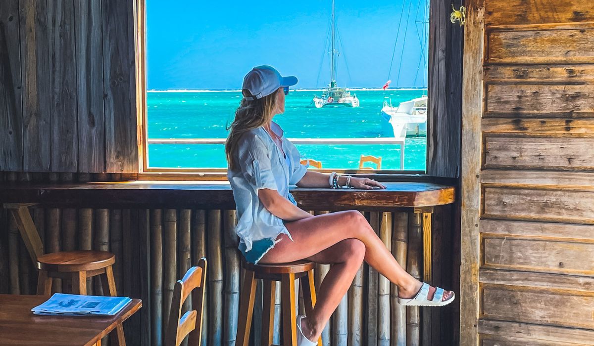 Kate sits on a tall wooden stool inside a rustic beachfront bar, gazing out through an open window at the turquoise ocean and sailboats in the distance. She wears a casual outfit with a white shirt, shorts, sandals, and a cap, embodying a relaxed, tropical vibe.