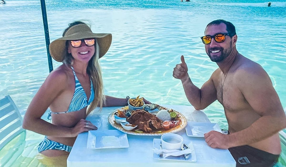 Kate and her husband sit at a table in clear blue water, enjoying a seafood feast in Belize. The man gives a thumbs-up while both wear sunglasses and smile at the camera. The large platter in front of them is filled with various Belizean foods, including fried fish, rice, and fries, showcasing local cuisine.