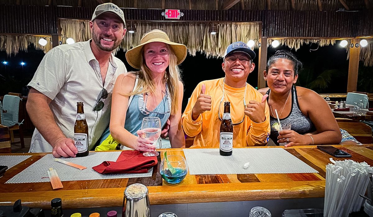 Four people smile and pose together at a bar in Belize, enjoying drinks and a relaxed evening. The group includes two men and two women, with one of the men giving a thumbs-up. They have a mix of beverages, including bottles of Belikin beer and cocktails.