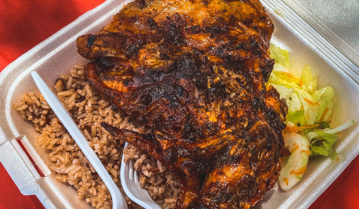 A takeout container filled with jerk chicken, served alongside beans and rice, and a small side of lettuce salad. The chicken is richly seasoned and charred, highlighting the bold and spicy flavors typical of jerk seasoning. The dish, paired with the classic Belizean beans and rice, offers a hearty and flavorful meal