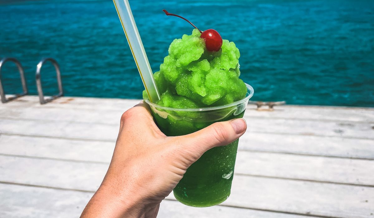 A hand holds a vibrant green frozen drink known as "Lizard Juice," topped with a cherry and served in a clear plastic cup with a straw. The refreshing beverage is enjoyed by the water, with the turquoise ocean and a wooden dock in the background.