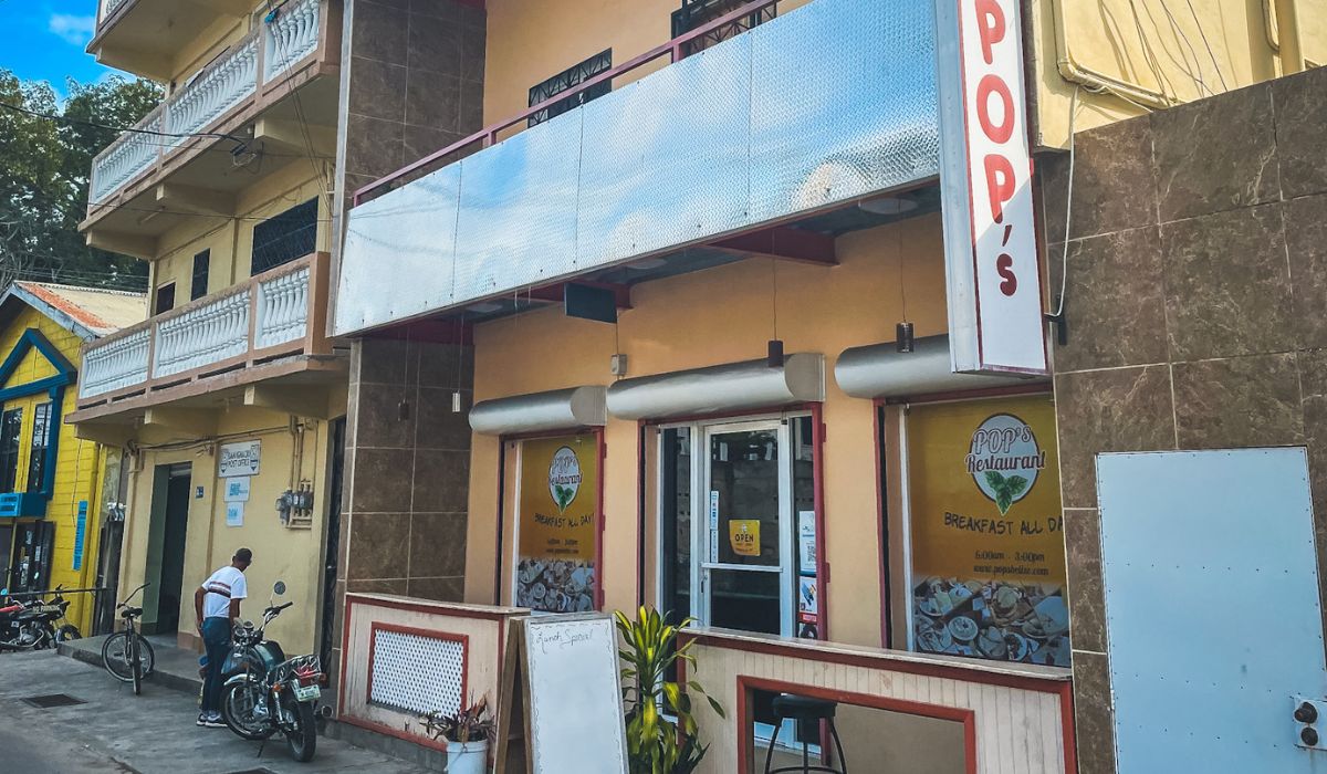 The exterior of Pop's Restaurant, a small, cozy eatery in Belize known for serving breakfast all day. The building has a simple, modest design with a bright sign reading "Pop's" above the entrance. A few motorcycles and bicycles are parked outside, while a handwritten chalkboard sign stands near the door, inviting customers in.