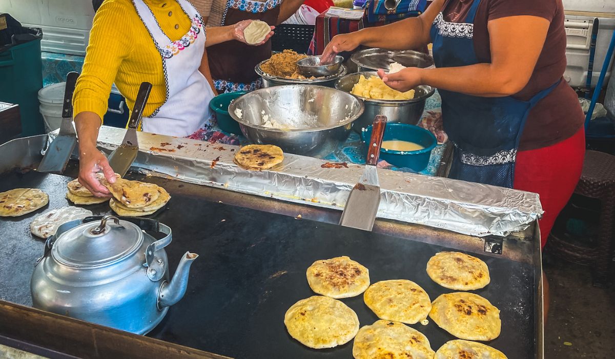 Women preparing and cooking pupusas at a food stall in Belize. The women are shaping the dough and stuffing it with various fillings, then cooking the pupusas on a large griddle. The scene showcases the hands-on, traditional process of making this popular dish, with bowls of ingredients and cooking utensils arranged around the workspace.