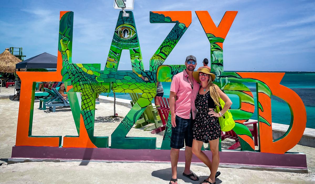 Kate and her husband pose in front of the colorful "Lazy Lizard" sign at a popular beach bar in Belize. The sign features a vibrant lizard design, with large letters that spell out "LAZY LIZARD," and is set against the backdrop of the turquoise ocean. The couple, dressed in casual summer attire, stands smiling under the bright sun.