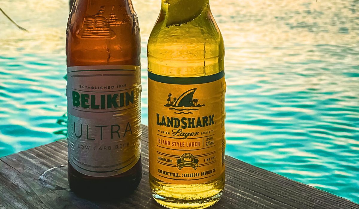 Two bottles of beer, Belikin Ultra and Landshark Lager, sit on a wooden surface with a backdrop of clear, turquoise water in Belize. The Belikin Ultra, a low-carb beer, represents the local Belizean brewery.