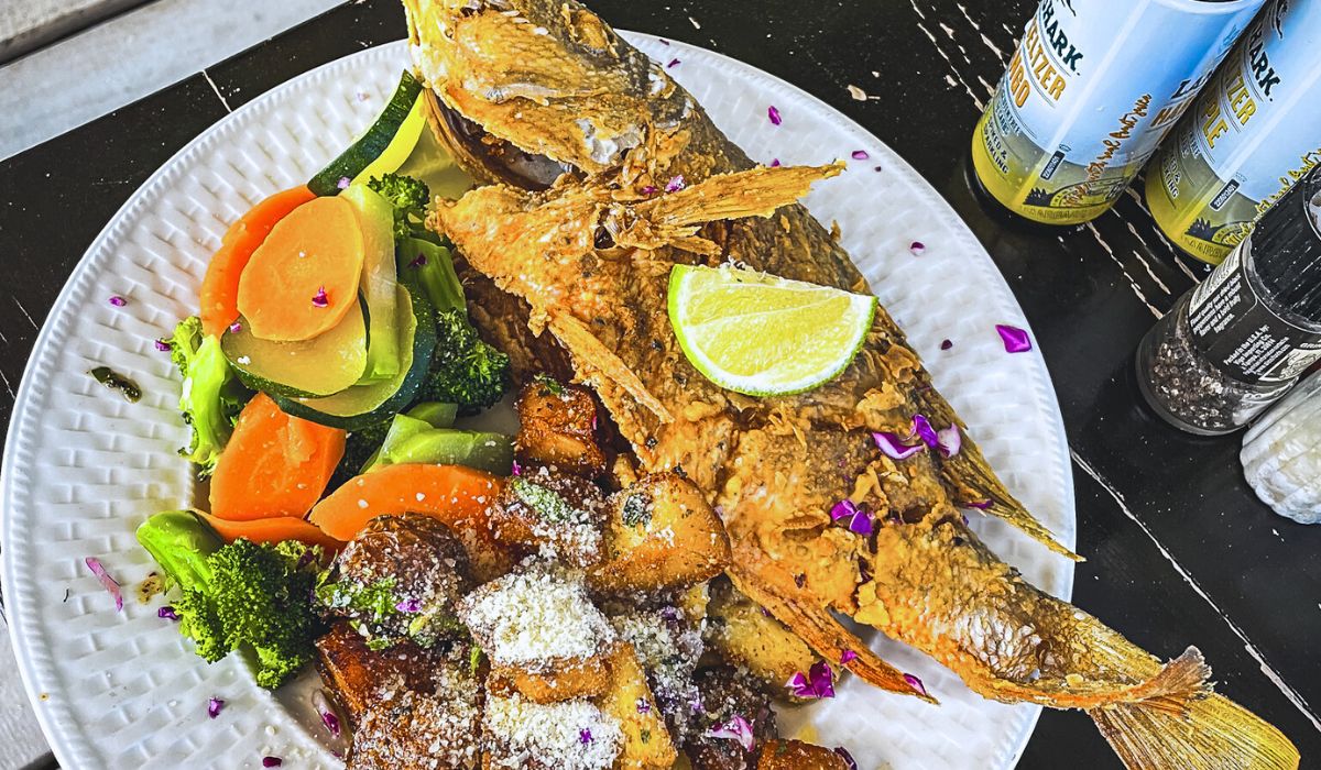 A plate featuring a whole fried snapper served with roasted potatoes and steamed vegetables, including broccoli, carrots, and zucchini. The snapper is garnished with a lime wedge and edible flower petals, adding a touch of color and freshness.