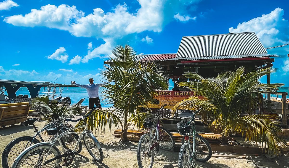 The Pelican Sunset Bar, a small beachside bar in Belize, is surrounded by palm trees and bicycles parked in the sandy area in front. A person stands with arms outstretched near the bar, enjoying the sunny weather and scenic ocean view. The bar’s rustic design with a corrugated metal roof and open-air seating creates a laid-back, tropical atmosphere perfect for relaxing by the water.