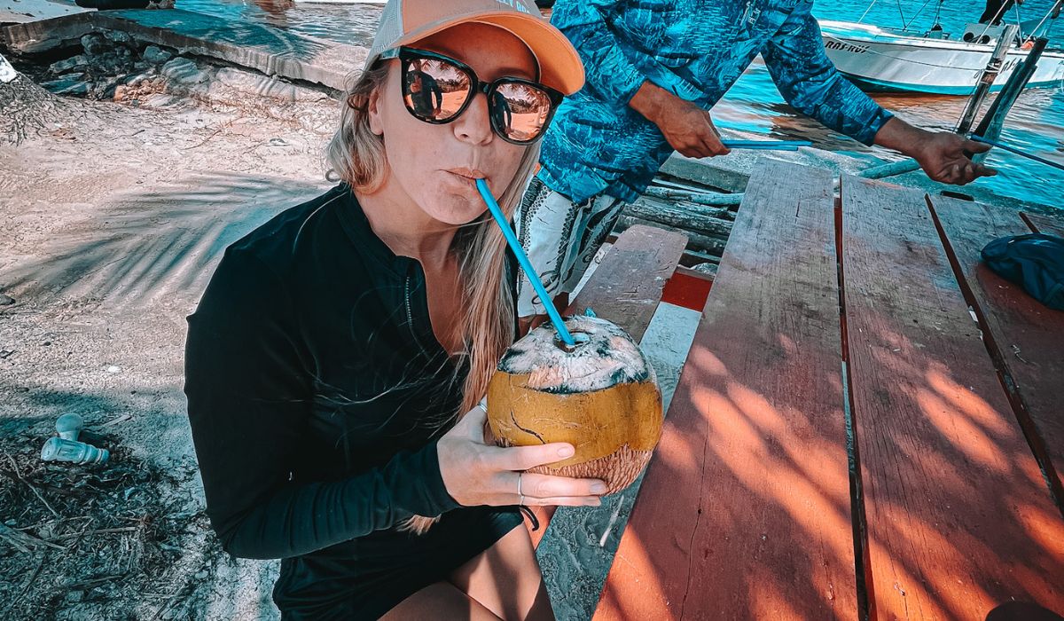 A woman wearing sunglasses and a cap sips from a fresh coconut with a straw while sitting at a picnic table near the water in Belize. The coconut has been cut open at the top, allowing access to the refreshing coconut water inside. The relaxed, tropical setting, with boats and the ocean in the background, highlights the laid-back vibe