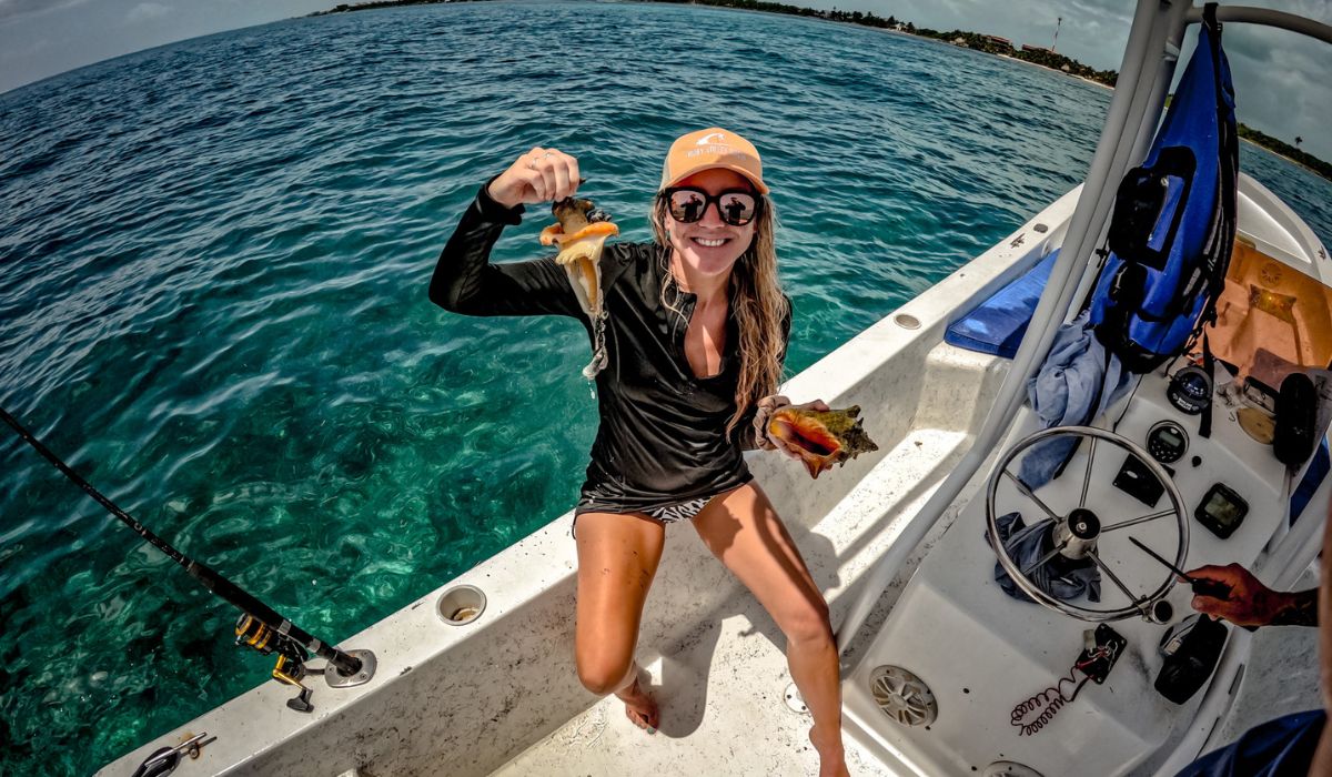 Kate, smiling and wearing sunglasses and a cap, holds up the freshly extracted meat from a conch shell while sitting on the edge of a boat in the clear blue waters of Belize.