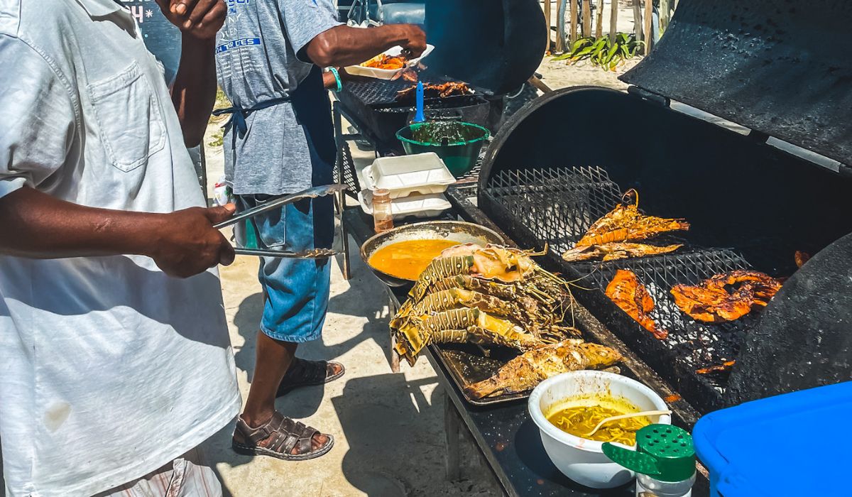 Two men grilling lobster tails and fish on large outdoor grills at a beachside barbecue in Belize. The scene shows a variety of seafood being prepared, with some pieces already cooked and others still grilling. Bowls of seasoning and sauce sit nearby, along with takeout containers ready to serve the freshly grilled food.