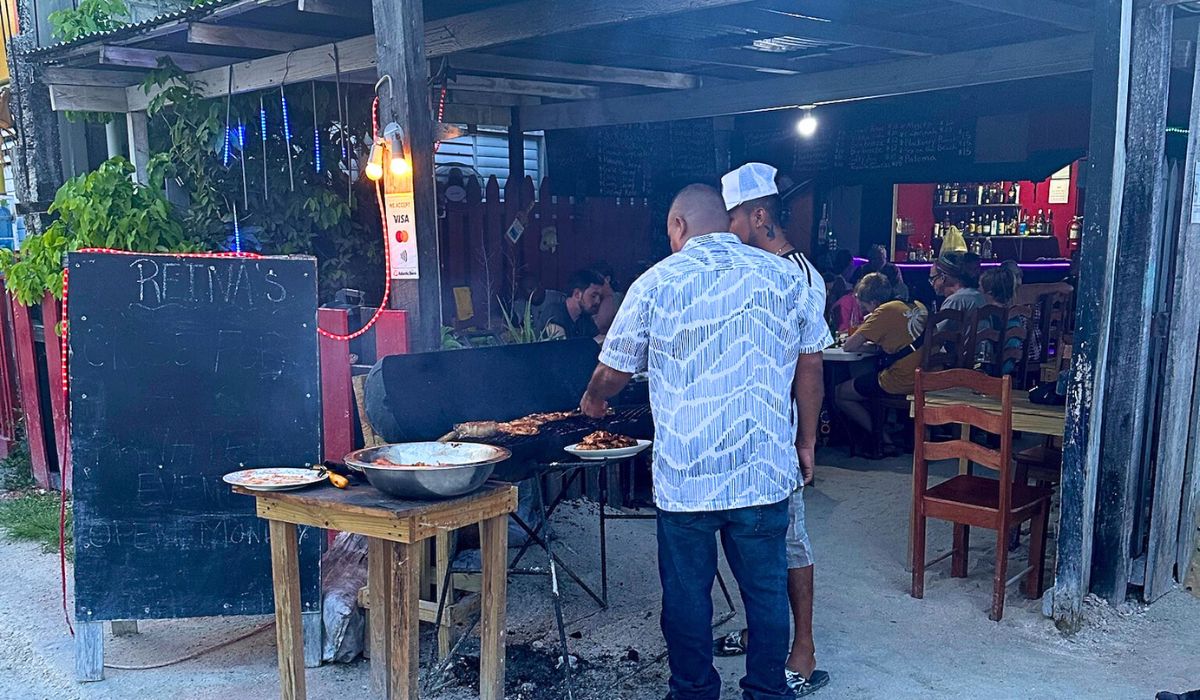 Two men grill food outside Reina's, a local restaurant in Belize, as evening approaches. The restaurant has an open, rustic setup with wooden furniture and a small bar inside, where patrons are seated and enjoying their meals. A chalkboard sign stands near the entrance.