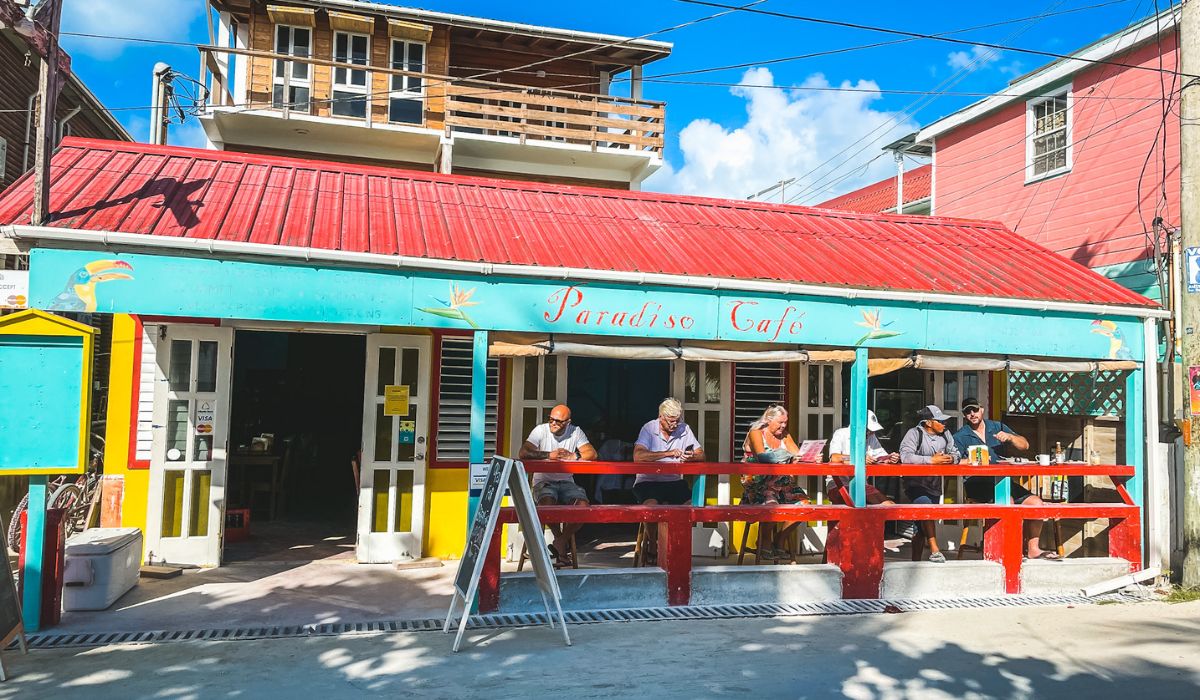 The exterior of Paradiso Cafe, a charming, colorful restaurant in Belize with a red roof and turquoise accents. The cafe has an open front with patrons sitting at the counter, enjoying their meals and drinks while looking out onto the street. The building's vibrant colors and casual seating area reflect the laid-back and welcoming atmosphere typical of local eateries in Belize.