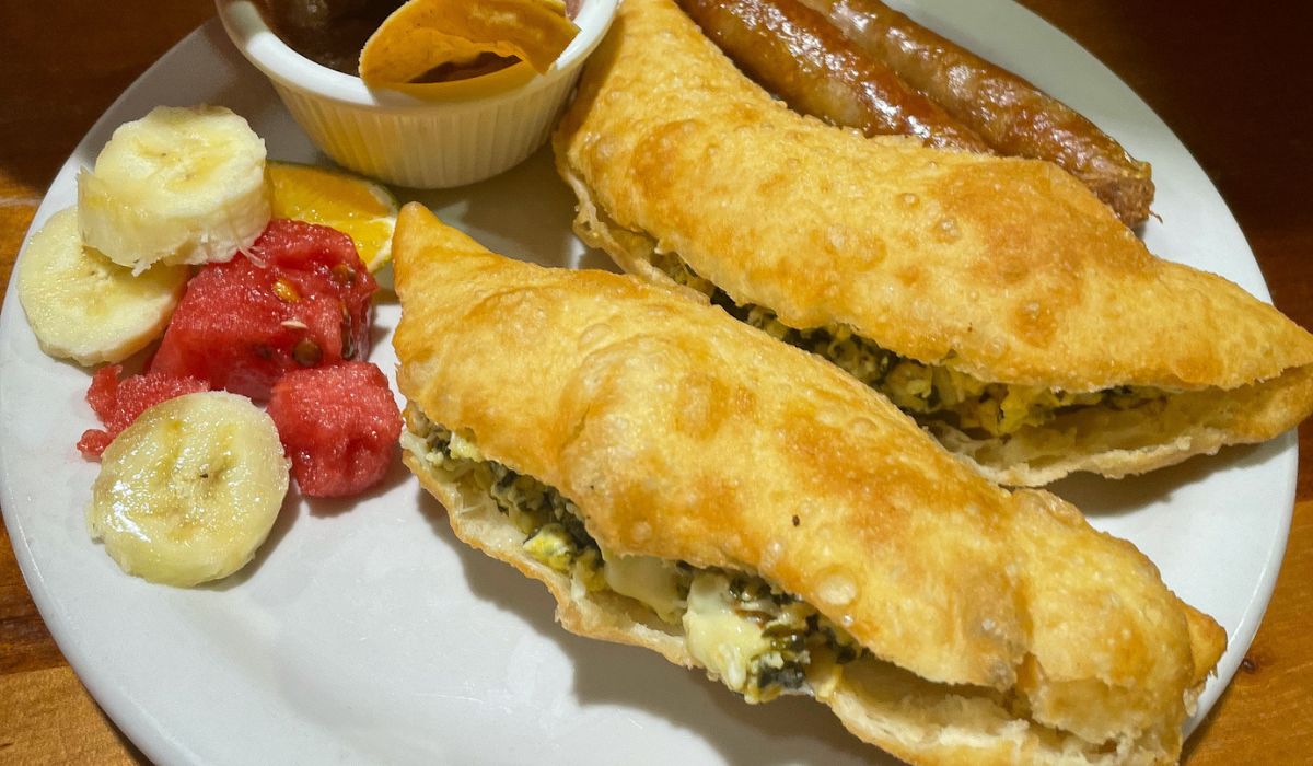 A plate of Belizean breakfast featuring two stuffed fry jacks filled with eggs and cheese, accompanied by sausage links, a small bowl of refried beans with tortilla chips, and a side of fresh fruit, including banana slices and watermelon. The fry jacks are golden and crispy, making them a quintessential part of Belizean cuisine.