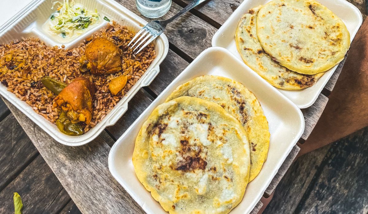 A meal featuring traditional Belizean beans and rice served with stewed chicken in a takeout container, accompanied by a side of slaw. Next to it, another container holds freshly cooked pupusas, stuffed with beans and cheese.