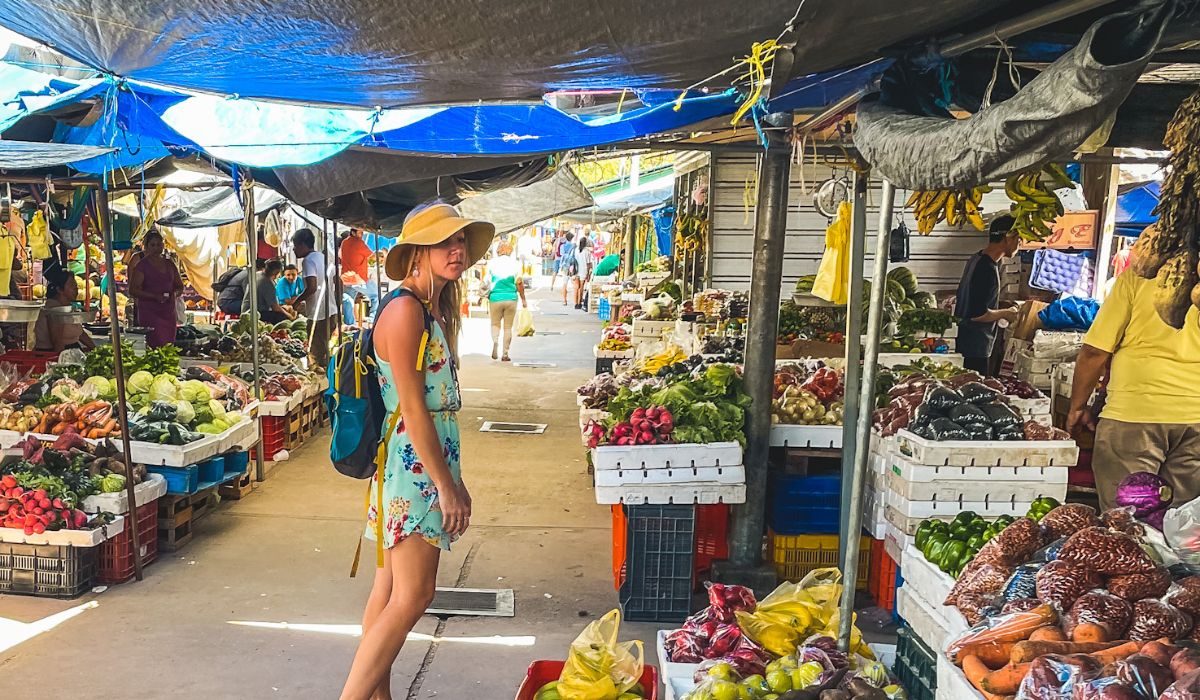  in a summer dress and wide-brimmed hat explores a vibrant outdoor market in Belize. She walks among stalls filled with fresh fruits, vegetables, and other produce, displayed in colorful piles under makeshift tarps. The market is bustling with activity as vendors and shoppers move through the narrow aisles.