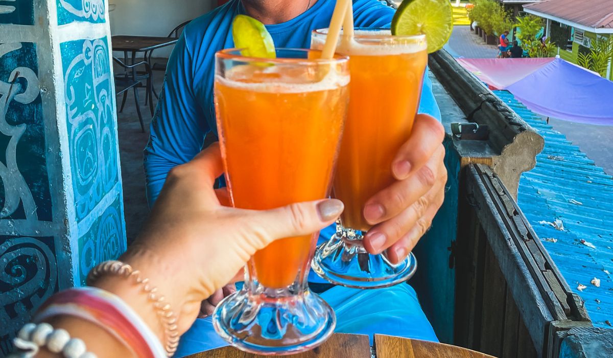 Two people raise glasses of vibrant orange Rum Punch, garnished with lime slices, while sitting at an outdoor bar in Belize. The glasses are held in a toast, capturing the celebratory and tropical vibe of enjoying a refreshing Rum Punch in a scenic setting. The background shows colorful rooftops and hints of a relaxed, island atmosphere.