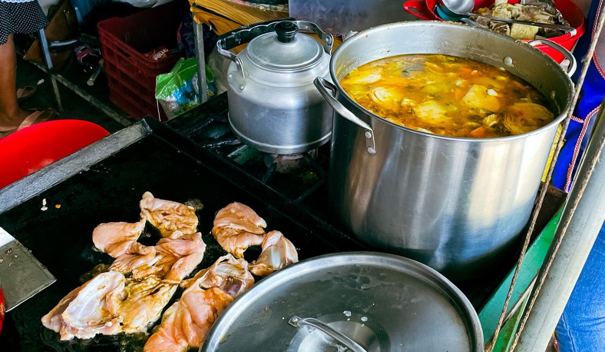 A large pot of stewed chicken simmers on a stove at a street food stall in Belize. The stew is rich with chicken pieces and vegetables, creating a hearty and flavorful broth. Next to the pot, pieces of marinated chicken are being grilled.