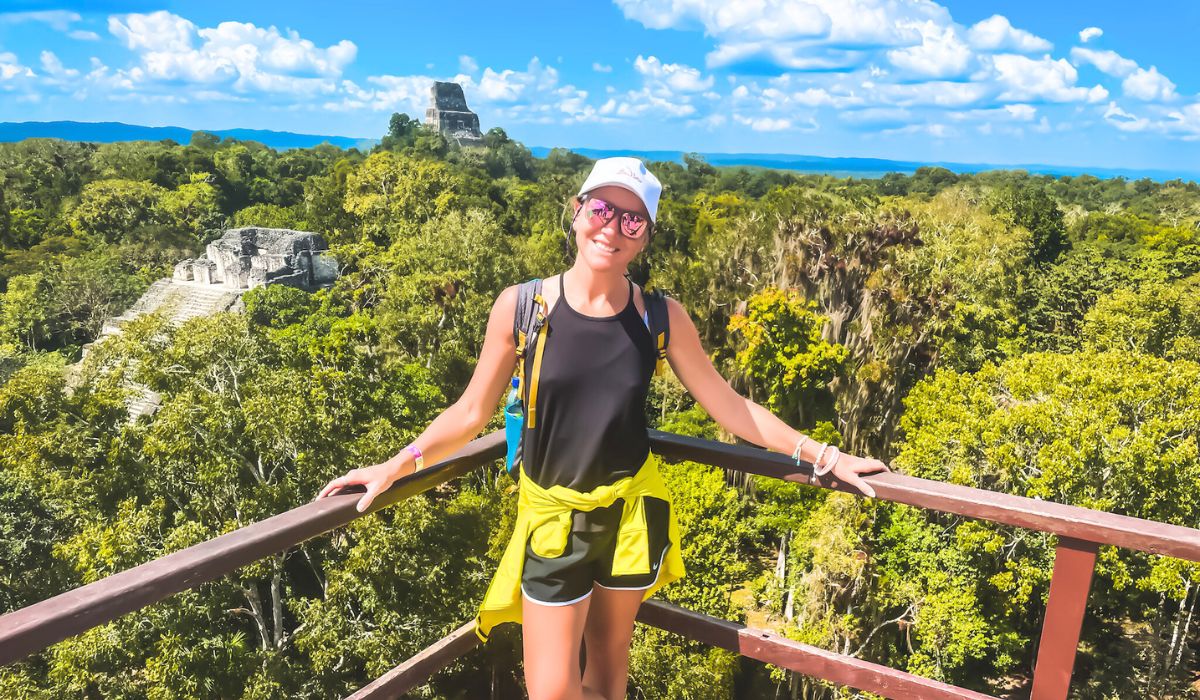 Kate, wearing a black tank top, shorts, and a white cap, poses on a wooden lookout point with lush Belize jungle and ancient Mayan ruins in the background under a bright blue sky. Demonstrating the amazing Belize Jungle Tours available.