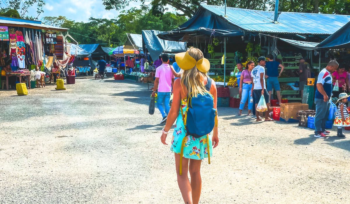 Kate, wearing a bright yellow hat, a floral dress, and a blue backpack is walking through a busy San Ignacio outdoor market. The market is filled with colorful stalls selling a variety of goods, including textiles, hammocks, and fresh produce. Shoppers and vendors are scattered throughout the scene, creating a lively atmosphere under the blue sky and surrounded by lush greenery.