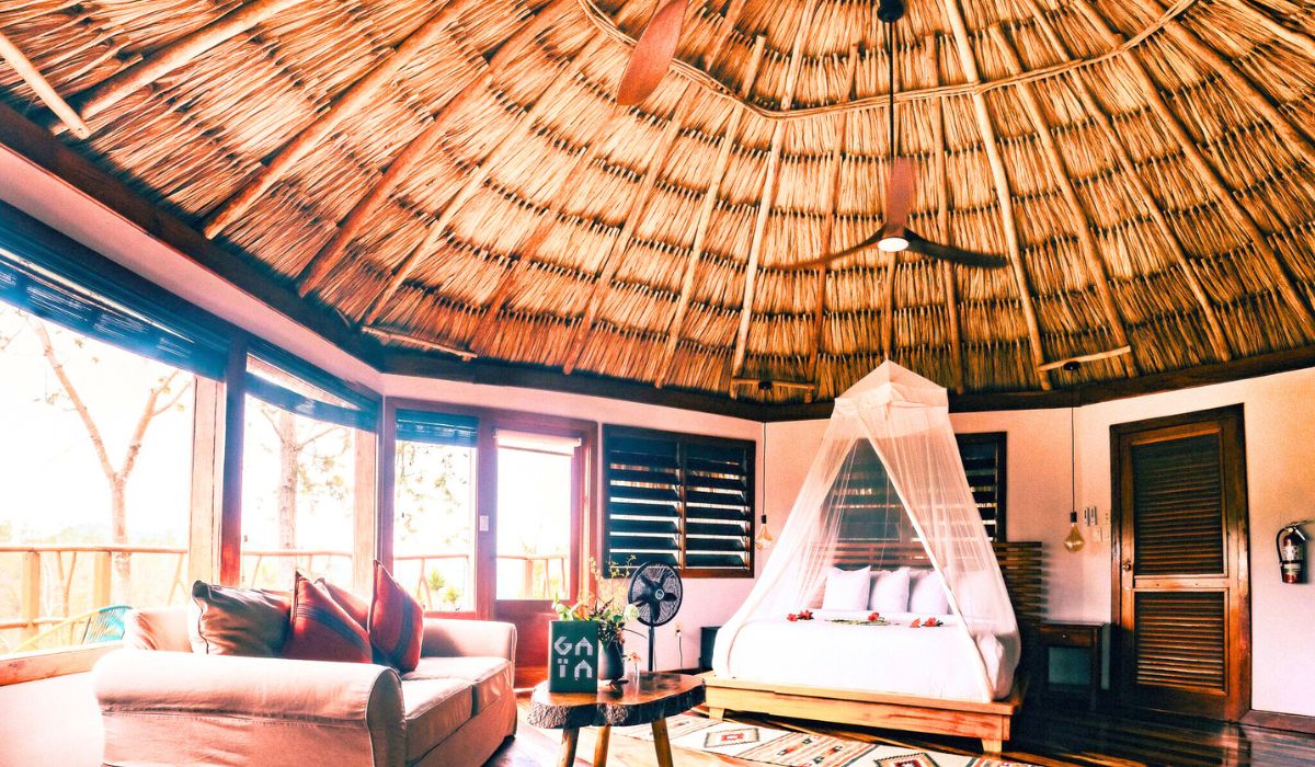 A cozy, round room with a thatched roof and a tropical ambiance. The room features a large bed draped with a mosquito net, a comfortable sofa with colorful pillows, and wooden furniture that complements the natural aesthetic. Large windows and doors allow for plenty of natural light and provide views of the surrounding jungle.