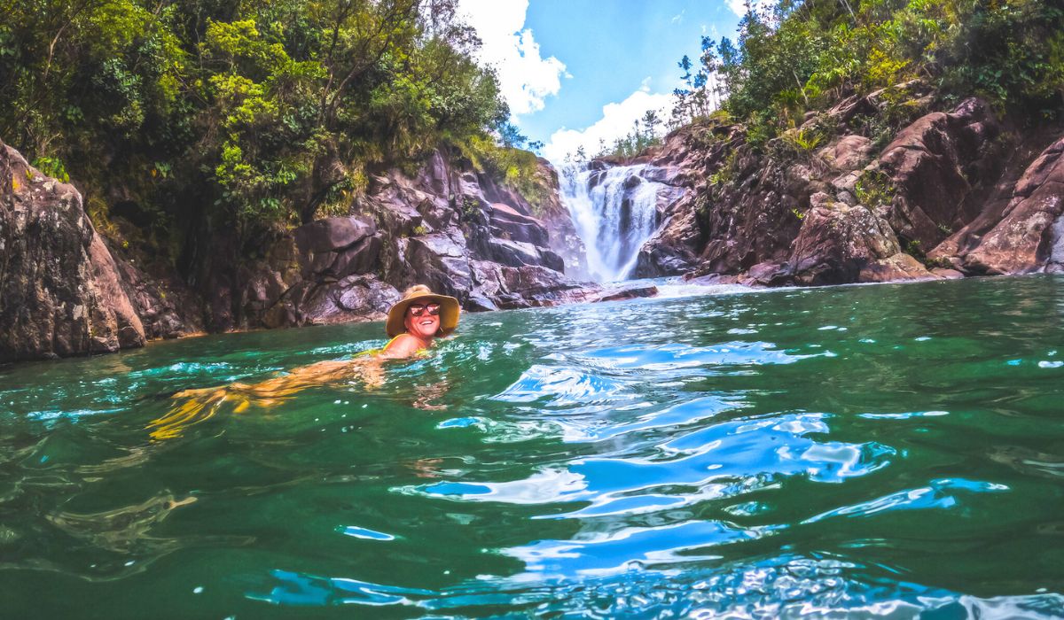 Kate, wearing a wide-brimmed hat, is swimming in a clear, greenish-blue natural pool, smiling at the camera. The pool is surrounded by rocky cliffs and lush vegetation, with a picturesque waterfall cascading in the background. The scene is serene and refreshing, capturing the joy of swimming in a secluded natural setting under a bright blue sky.