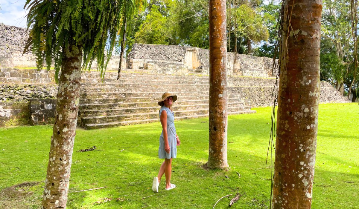 Kate, wearing a light dress and a wide-brimmed hat stands in a grassy area in front of ancient stone steps, part of a Mayan ruin site. Tall trees frame the scene, providing a mix of shade and sunlight. The stone structures in the background contrast with the vibrant greenery, creating a peaceful, historical setting. Kate is looking back over her shoulder.