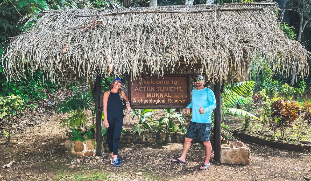 Kate and her husband are standing under a thatched-roof entrance sign for "Actun Tunichil Muknal Archaeological Reserve" in a lush, tropical setting. Kate is wearing black athletic wear and a cap, while her husband is dressed in a light blue long-sleeve shirt and shorts, giving a thumbs-up.