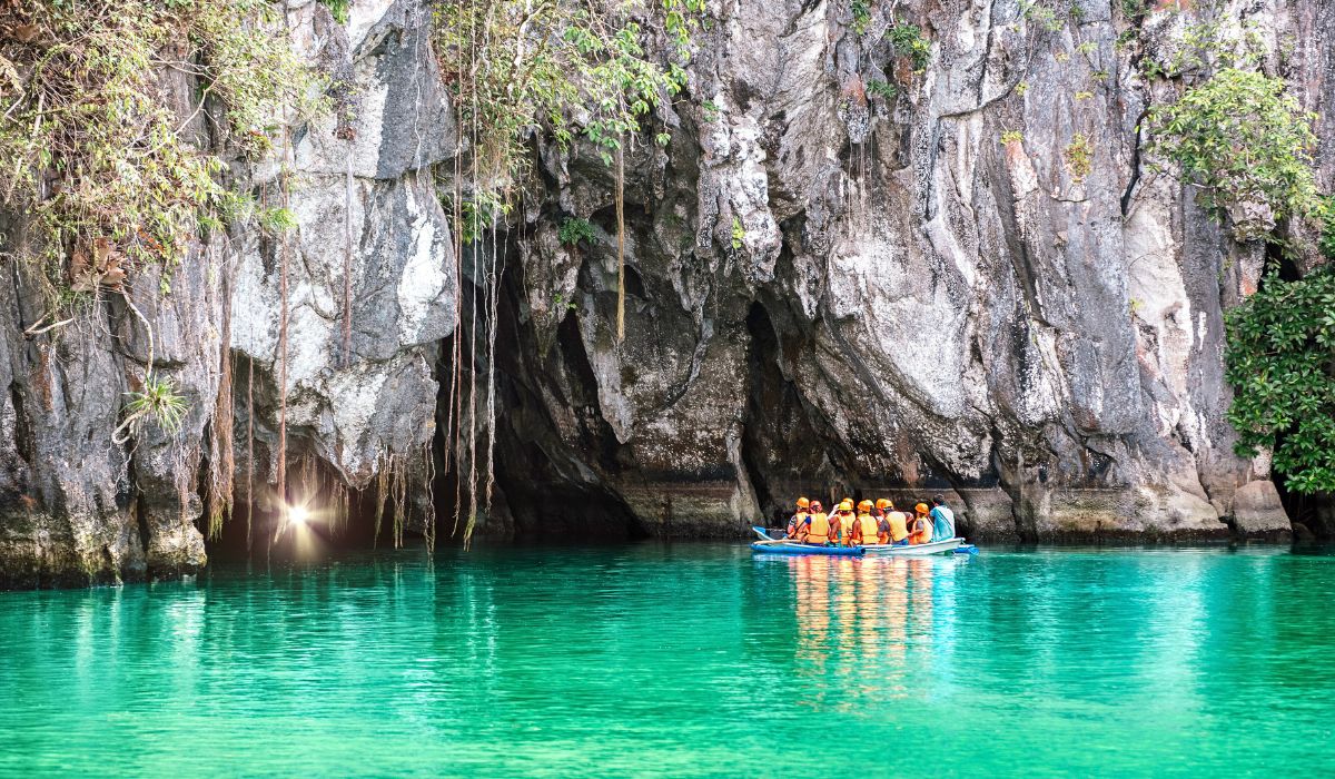 A group of people wearing orange life jackets and helmets are in a small boat approaching the entrance of a cave with rugged rock walls and hanging vegetation. The turquoise water is calm, reflecting the cave and the surrounding greenery. A soft light glows from within the cave entrance, adding a sense of mystery to the scene.