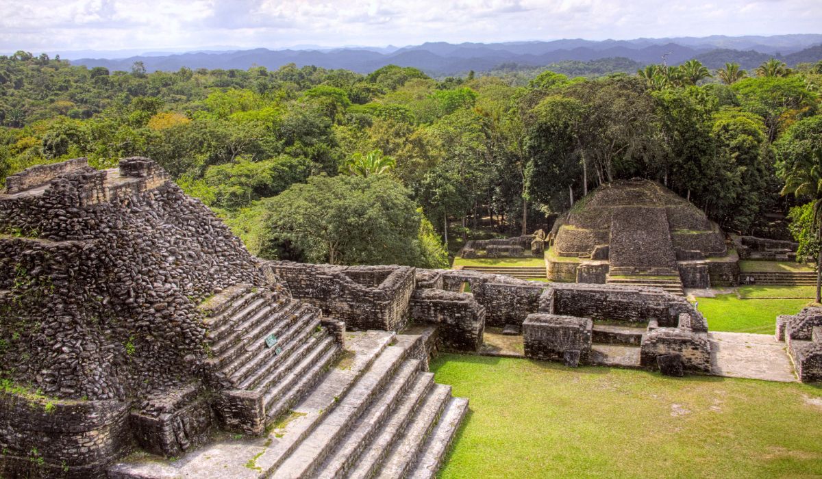 An expansive view of ancient Mayan ruins set against a backdrop of dense jungle and rolling hills. The stone structures include large pyramidal temples with staircases leading to the top and surrounding platforms.