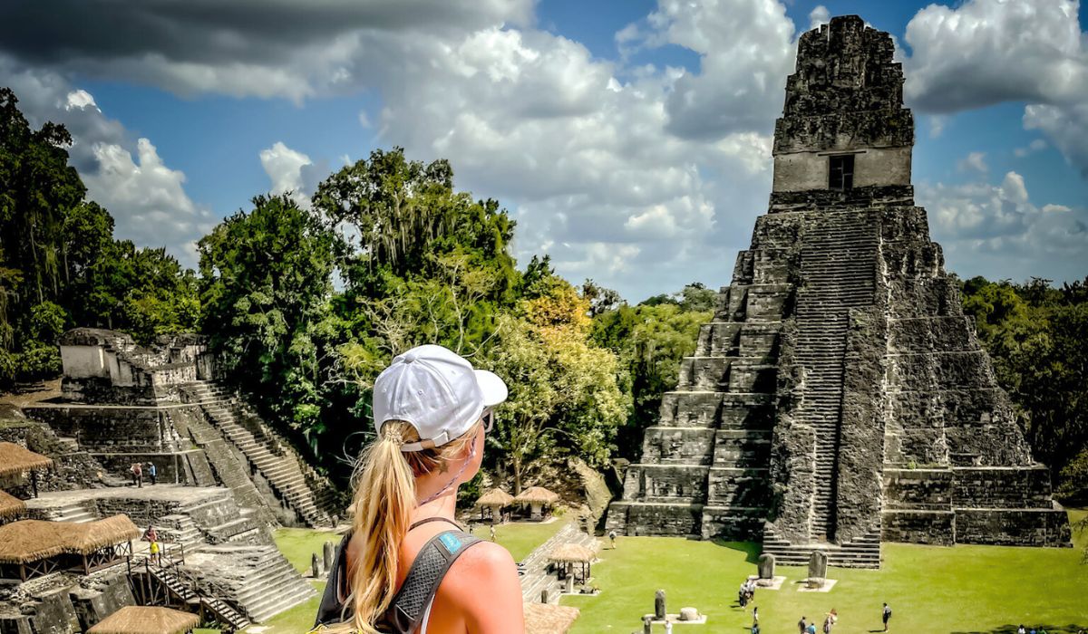 The image shows Kate with a white baseball cap and a backpack standing and looking at the iconic Temple I (also known as the Temple of the Great Jaguar) at Tikal, a major Mayan archaeological site in Guatemala. The temple is a tall, stepped pyramid with steep stone stairs, surrounded by lush jungle and other ancient structures.