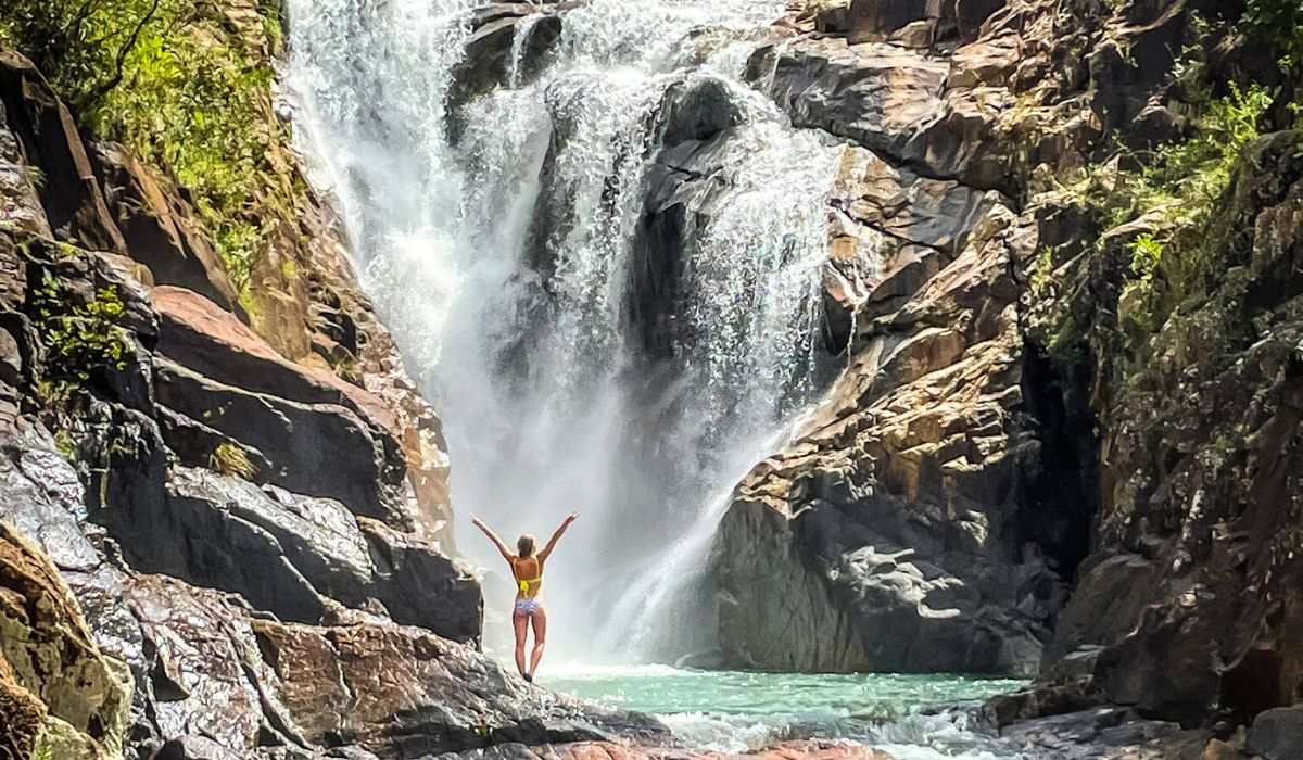 Kate in a yellow swimsuit stands with arms raised in front of a powerful waterfall cascading down rocky cliffs into a pool of water below. The scene captures the energy and majesty of the natural surroundings, with the waterfall creating a misty spray as it hits the rocks, and the person appearing small in comparison to the towering waterfall.