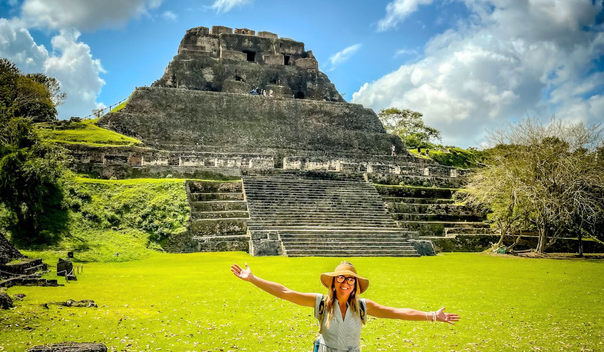 Kate, in a wide-brimmed hat and sunglasses, stands with arms outstretched in front of the large, stepped pyramid of Xunantunich, a Mayan archaeological site in Belize. The pyramid, known as "El Castillo," rises impressively in the background, with a wide staircase leading up to its summit. The lush green lawn and surrounding trees contrast with the ancient stone structure, and the clear blue sky adds to the vibrant atmosphere of the scene.