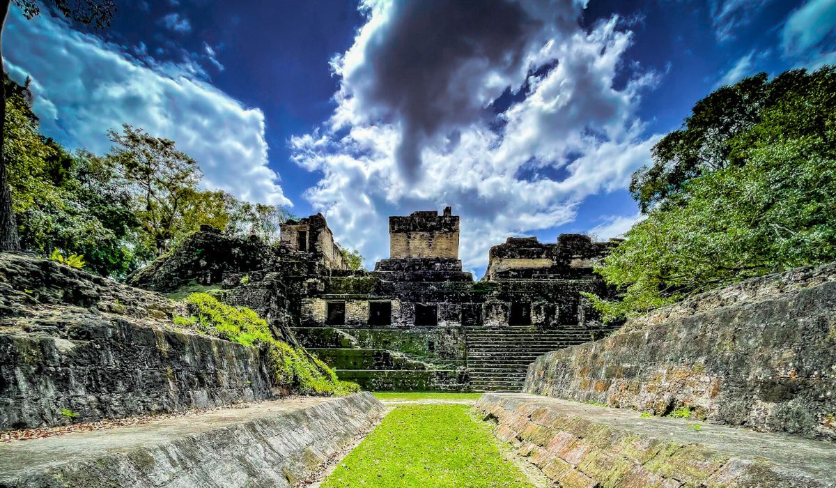 The image shows an ancient Mayan structure at Tikal, with a wide stone pathway leading up to a series of stepped terraces and buildings. The ruins are surrounded by lush greenery, and a dramatic sky with thick, rolling clouds looms overhead, adding a sense of mystery and grandeur to the scene.