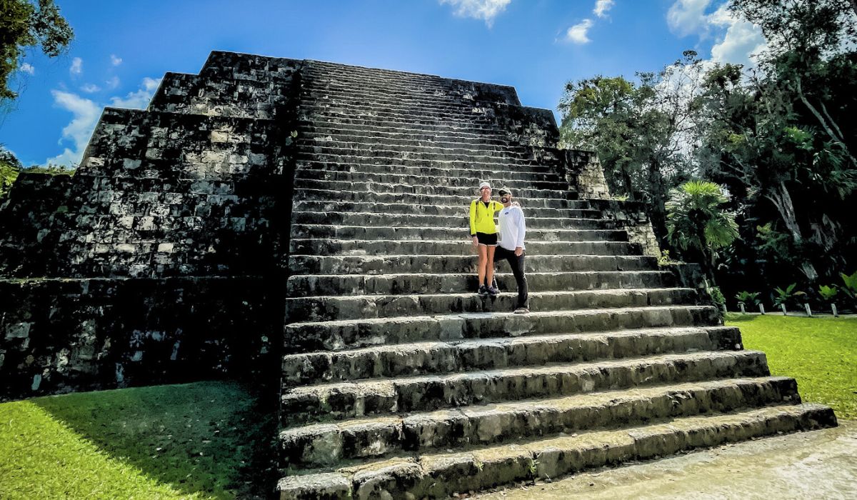 The image shows Kate and her husband standing on the wide stone steps of a large Mayan pyramid in Tikal, with the structure towering above them. They are positioned near the center of the pyramid, smiling and embracing, dressed in casual, athletic attire. The pyramid's steep, weathered steps rise dramatically against a bright blue sky, surrounded by lush greenery. 